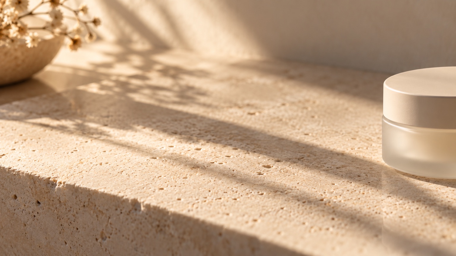Raking side light across a faintly dimpled pale stone tile, a plain frosted jar at the edge of the frame.