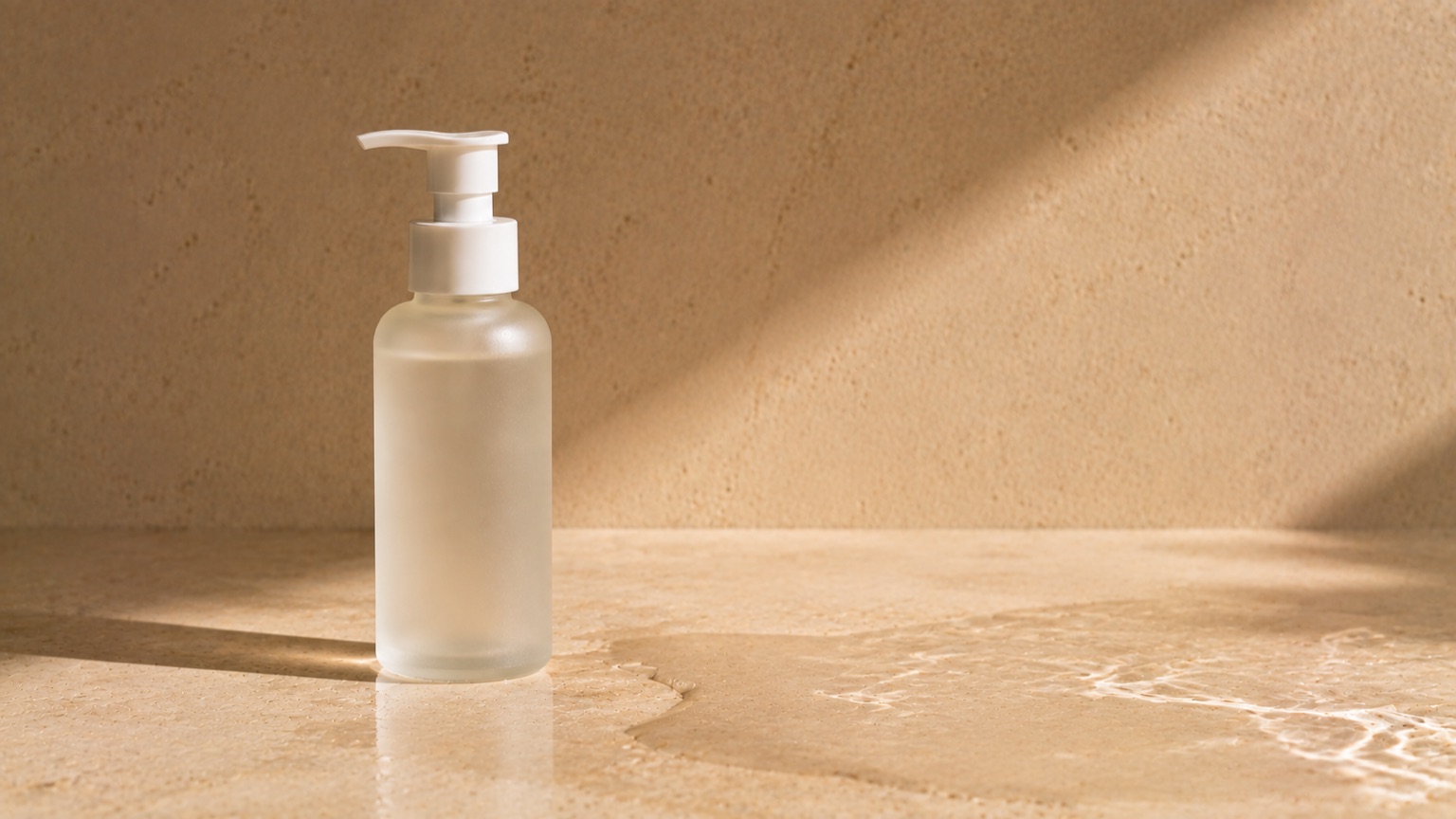 A plain frosted pump bottle standing on wet porcelain beside a shallow pool of water catching the light.