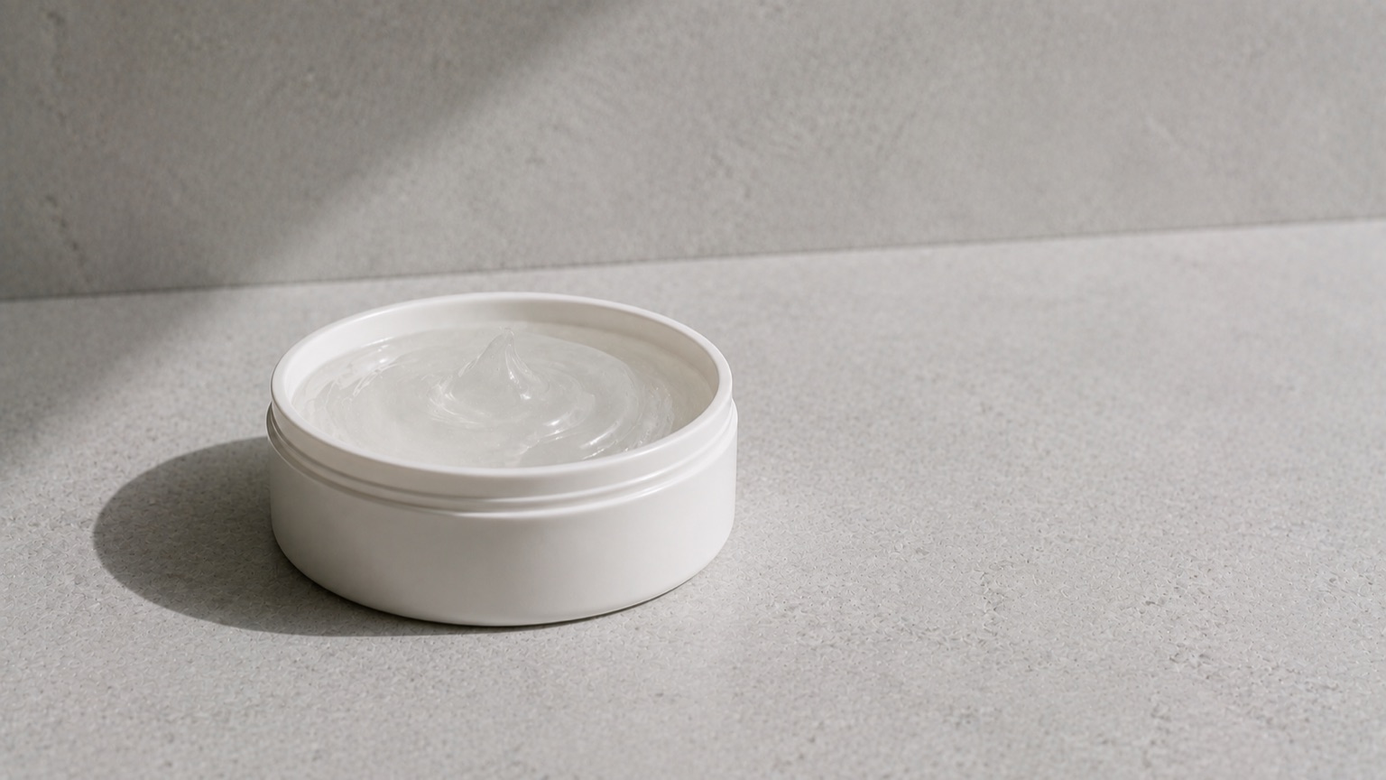 A shallow wide unlabelled jar on cool grey stone, a thin gel-textured swirl catching the light.