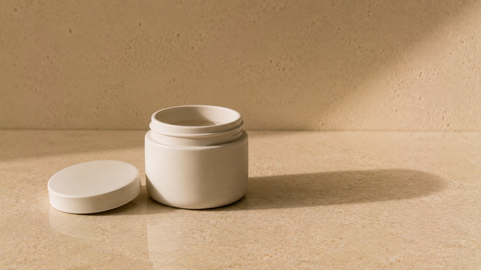 A plain matte ceramic jar with its lid set beside it on warm porcelain, casting a long soft shadow.