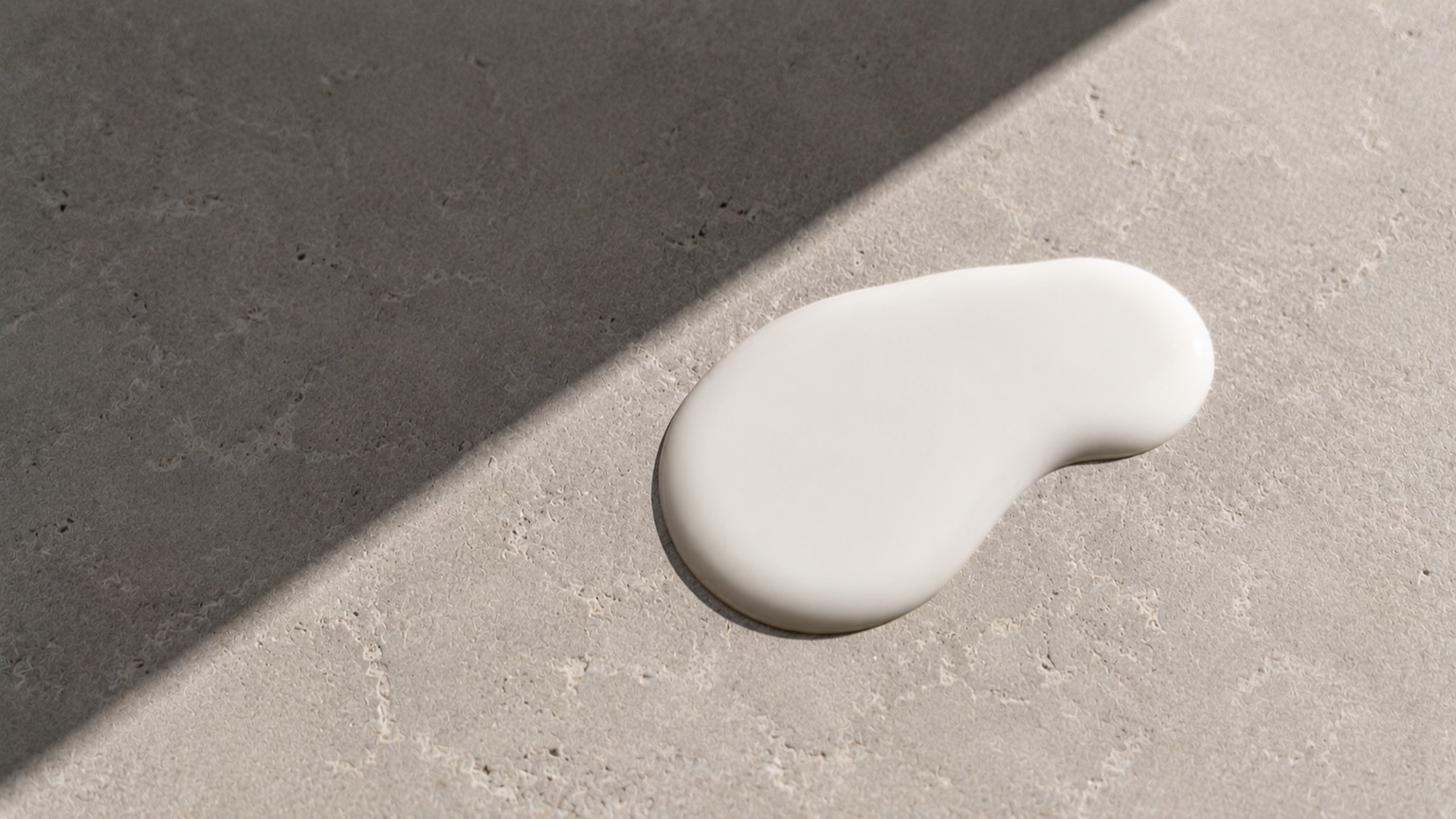 Matte white fluid pooled on textured grey stone under hard midday light, cut by one crisp geometric shadow.