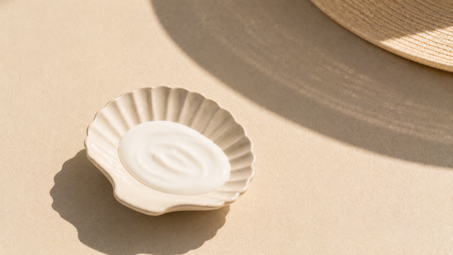 White lotion pooled in a shallow shell-shaped dish under strong midday sun, with the curved shadow of a hat brim across it.