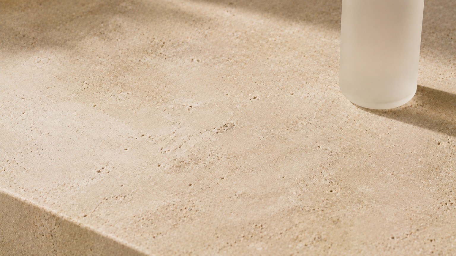 A pale stone slab with natural mineral speckling under raking light, a frosted bottle at the edge of frame.