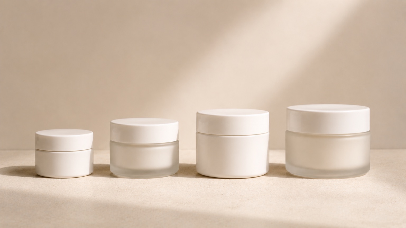 Four unlabelled white and frosted-glass moisturiser jars of increasing size lined up side by side.