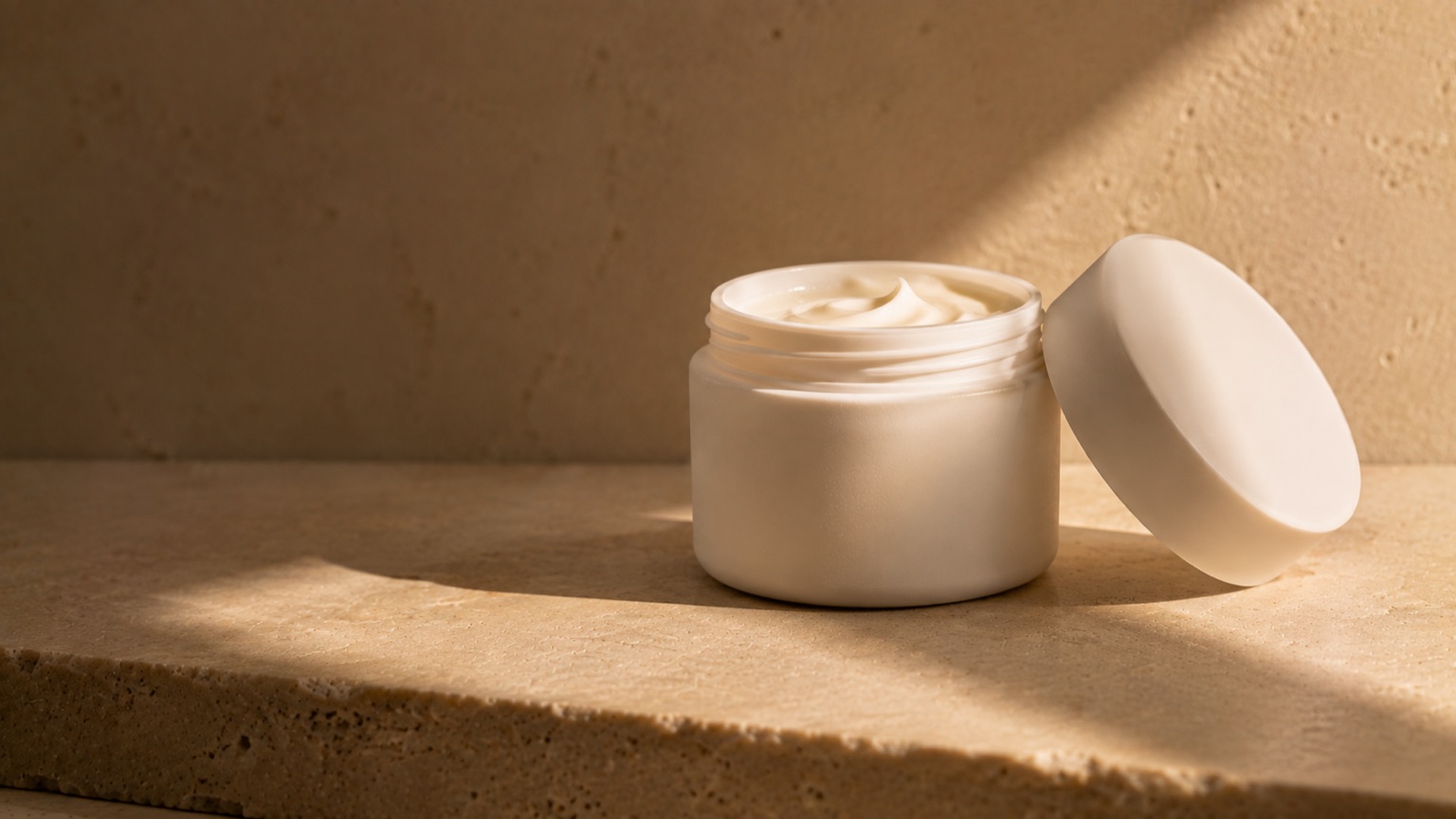 A plain matte white jar with its lid resting beside it, a soft swirl of cream visible at the rim.