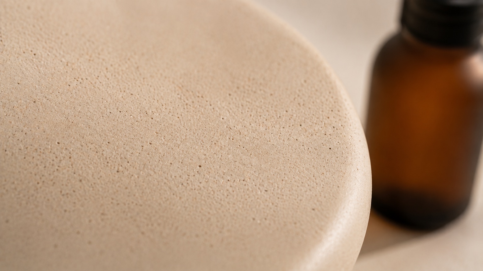 A macro view of an unglazed ceramic surface showing fine natural porosity, a plain glass bottle blurred at the edge.