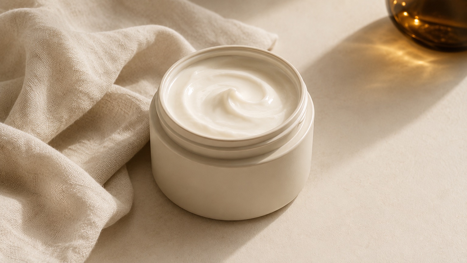 An open ceramic jar of thick cream with a soft swirl on its surface, resting on crumpled pale linen.