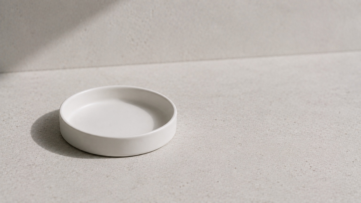 An empty plain white ceramic dish on pale stone in cool restrained daylight, holding nothing.